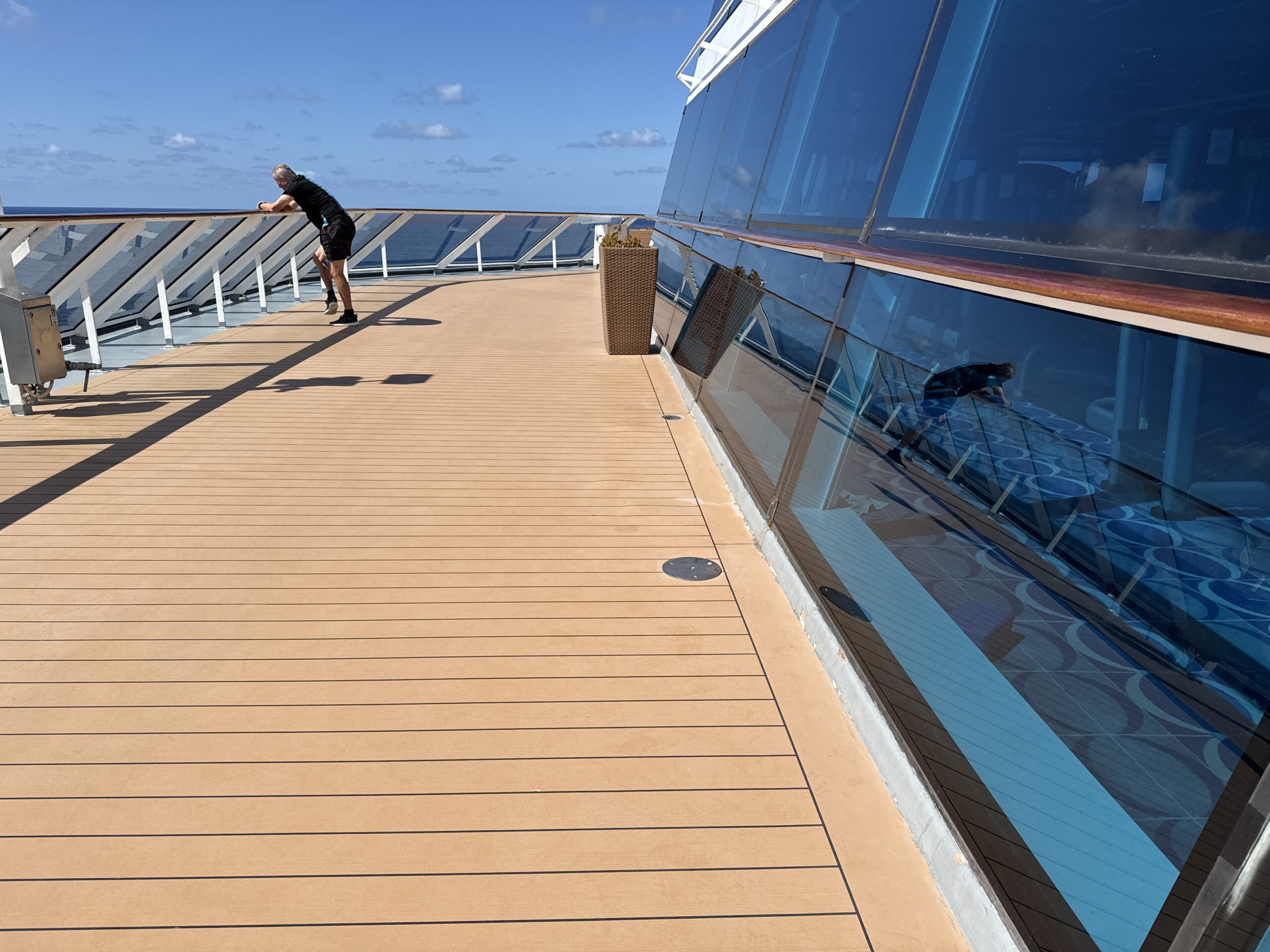 A man leans on the glass railing of a sunny teak deck aboard Celebrity Solstice, open ocean beyond.