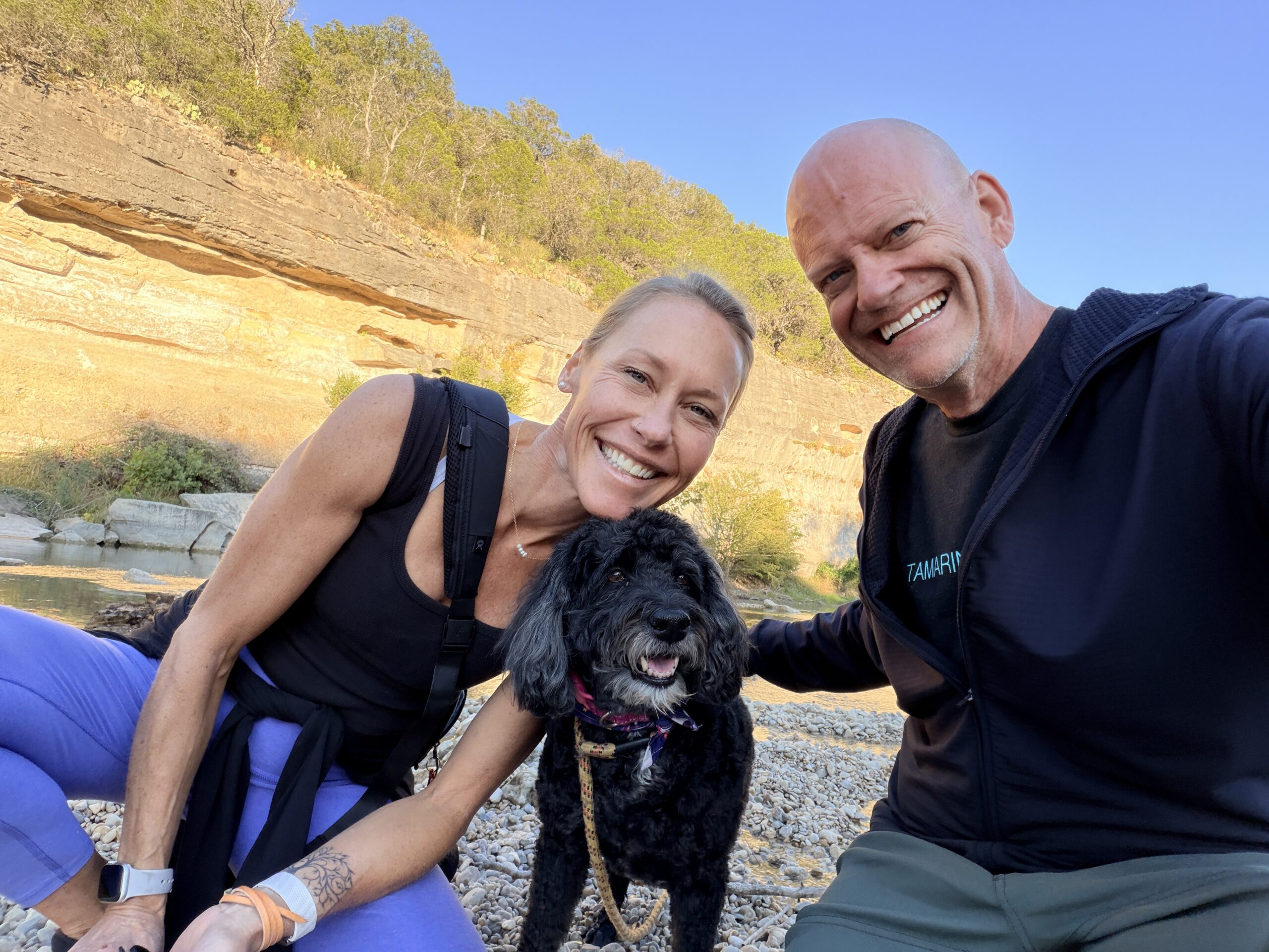 Matt and Erin smiling on a rocky riverbank with Rindo, their senior dog, below a limestone bluff.