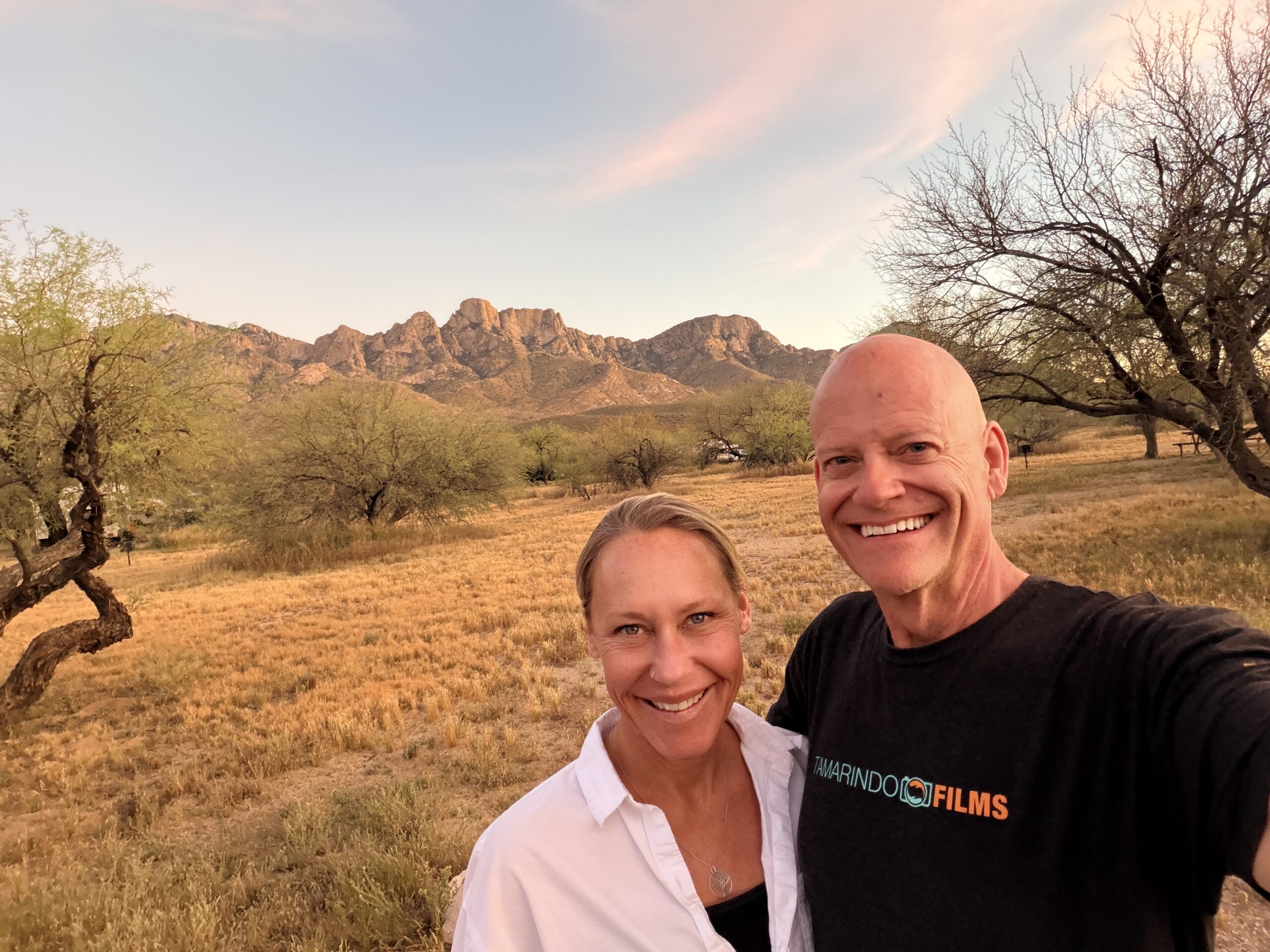 Matt and Erin taking a selfie in a grassy desert campground with rugged mountains behind them at dusk.