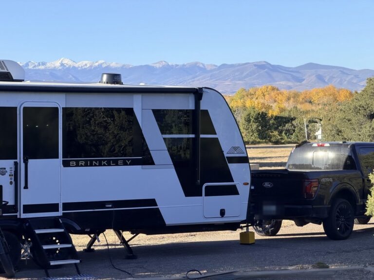 A white Brinkley travel trailer hitched to a black pickup at camp, with yellow fall trees and snowy mountains behind.