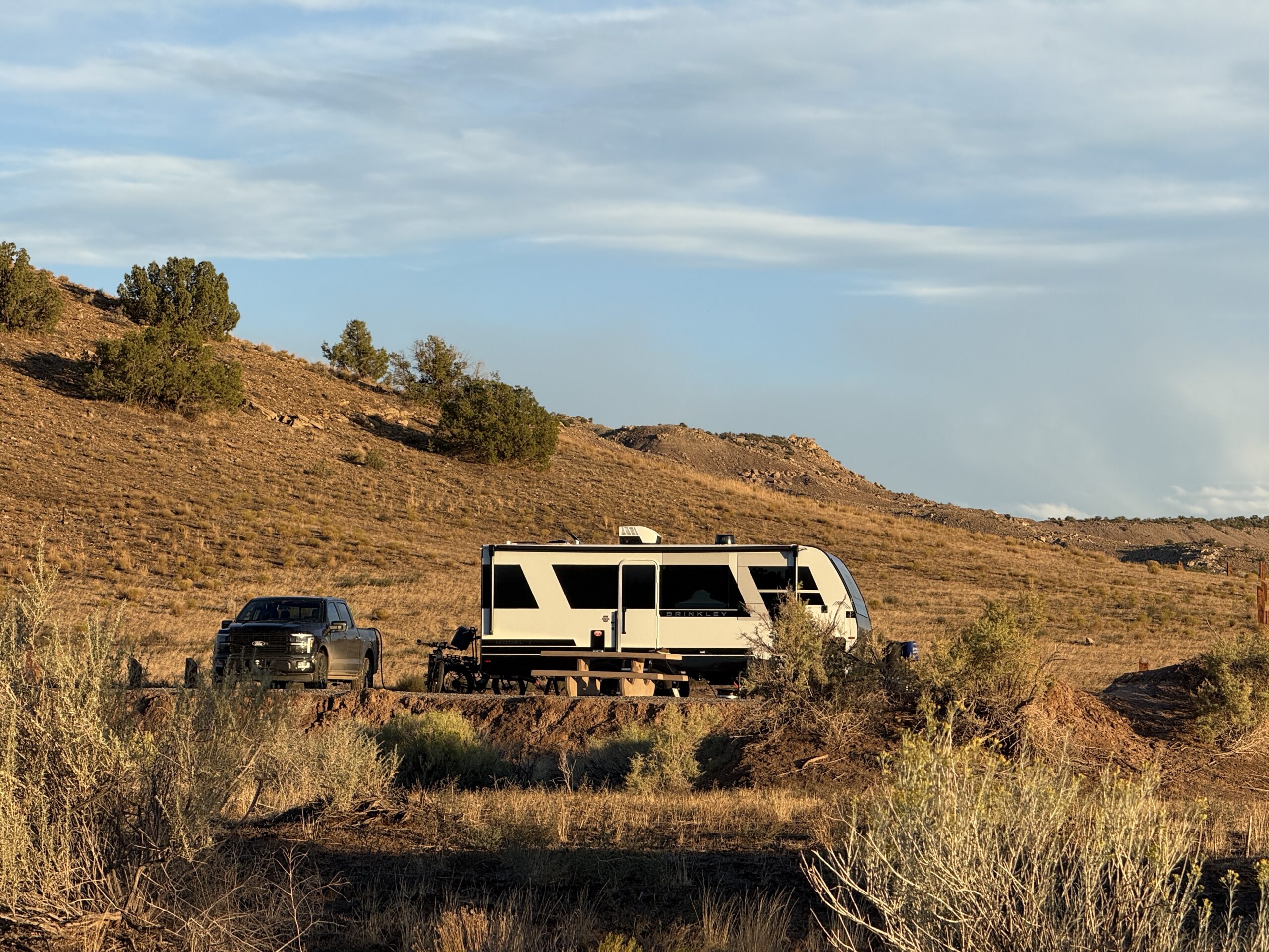 Brinkley trailer and pickup truck parked below a juniper-dotted hillside in Colorado in warm evening light.