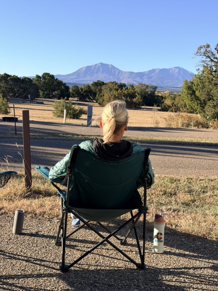 Erin sits in a green camp chair at a campsite, looking out across open grassland to a pair of mountain peaks.
