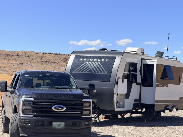 A black Ford pickup parked in front of the Brinkley travel trailer at a gravel desert campsite.