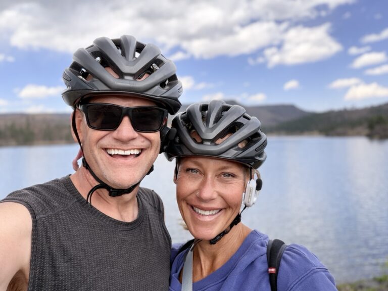 Matt and Erin smiling in bike helmets in front of a calm lake and forested hills.