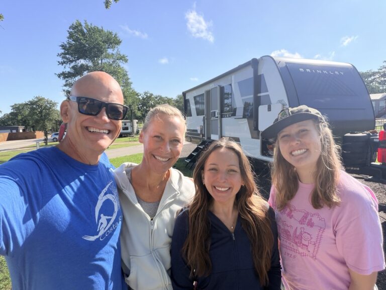Matt and Erin with their two daughters, all smiling, in front of the Brinkley trailer at a grassy campground.