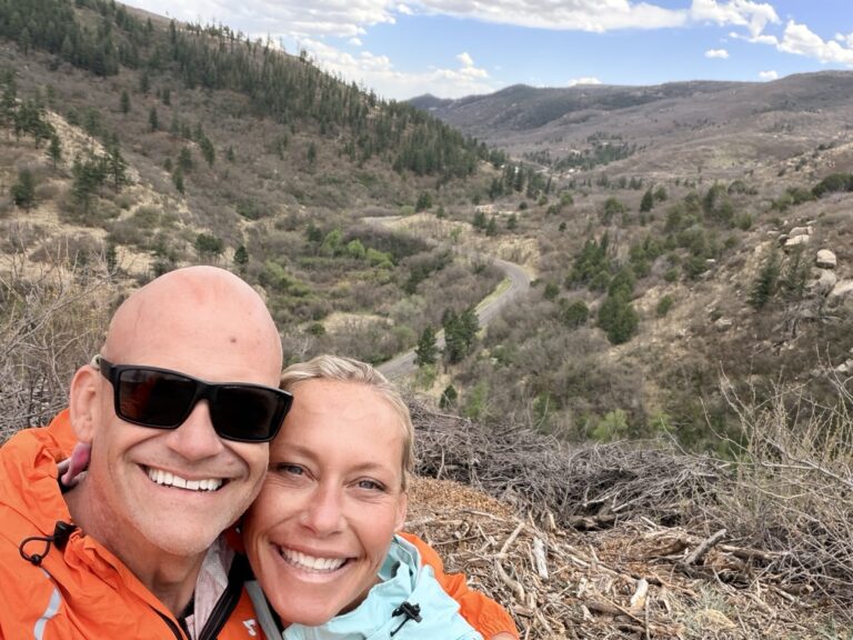 Matt and Erin in rain jackets smiling at an overlook above a winding road through a forested valley.