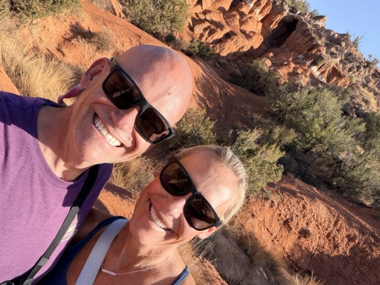 Matt and Erin in sunglasses taking a selfie on a red dirt trail below eroded red rock cliffs.