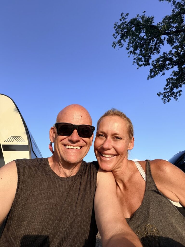 Matt and Erin smiling in a sunny selfie beside the front of their Brinkley trailer under a blue sky.