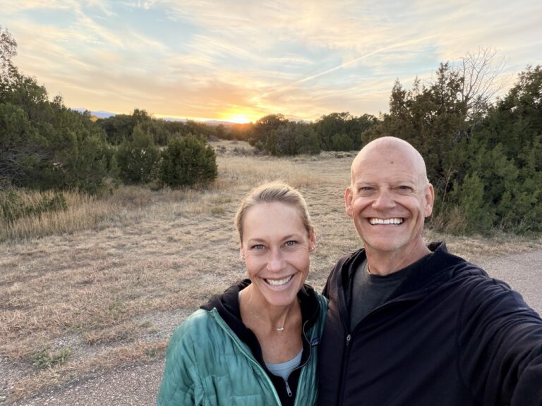 Matt and Erin smiling in a selfie on a dirt path as the sun sets behind juniper trees and dry grass.