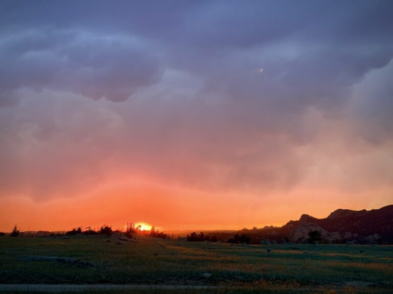 An orange sunset glowing beneath heavy storm clouds over a grassy plain with rocky outcrops on the horizon.