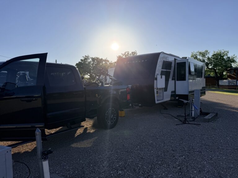 A black pickup hitched to the Brinkley travel trailer at a gravel RV site with the sun low in the sky.
