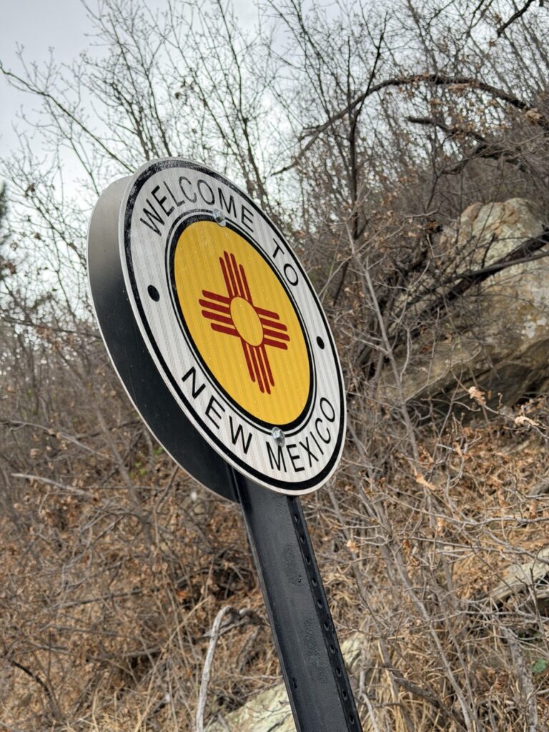 A round Welcome to New Mexico sign with the red Zia sun symbol on a yellow background beside a brushy hillside.