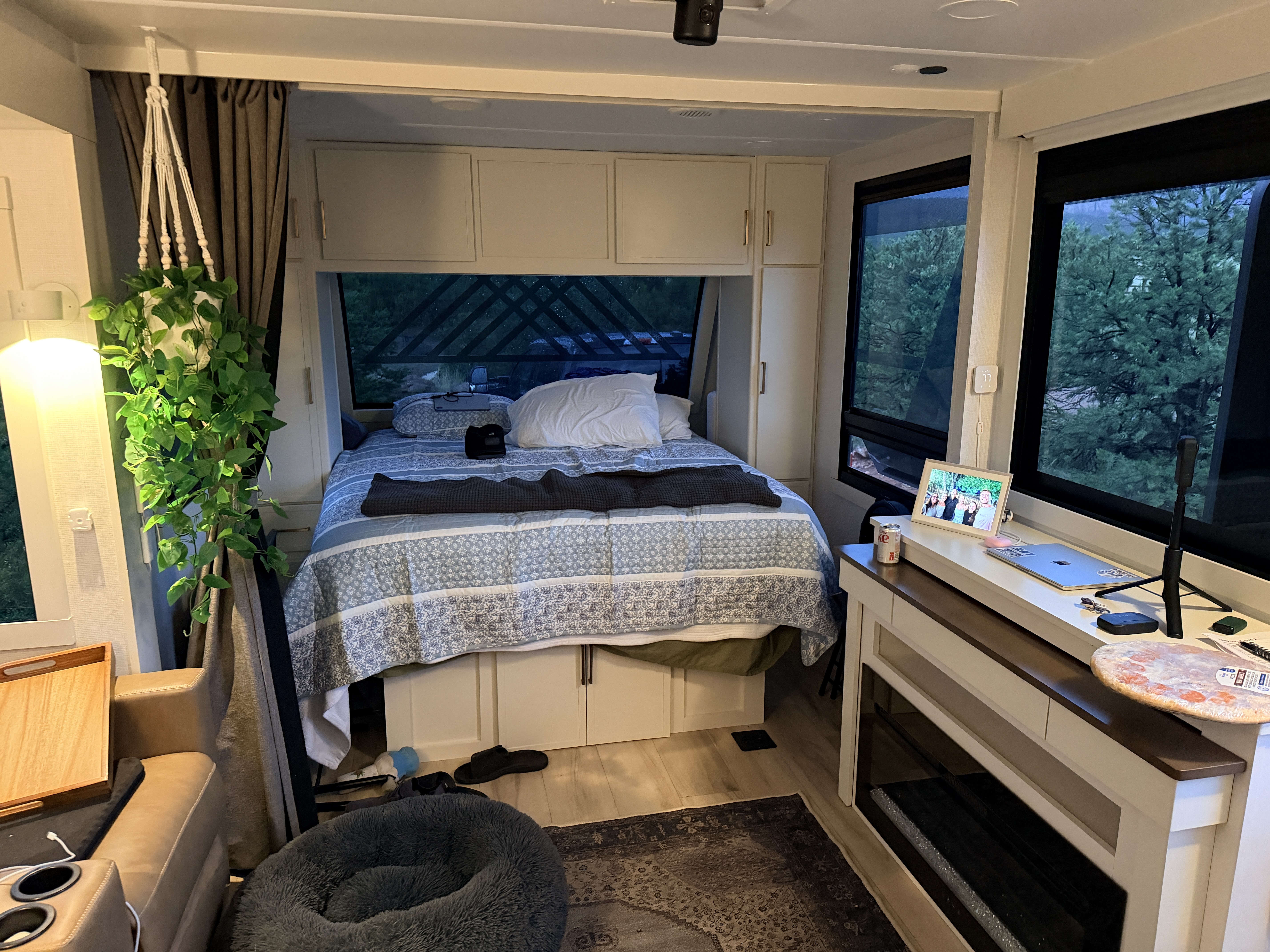 Interior of the Brinkley travel trailer looking toward the bed, with a hanging plant, desk and pine trees outside.