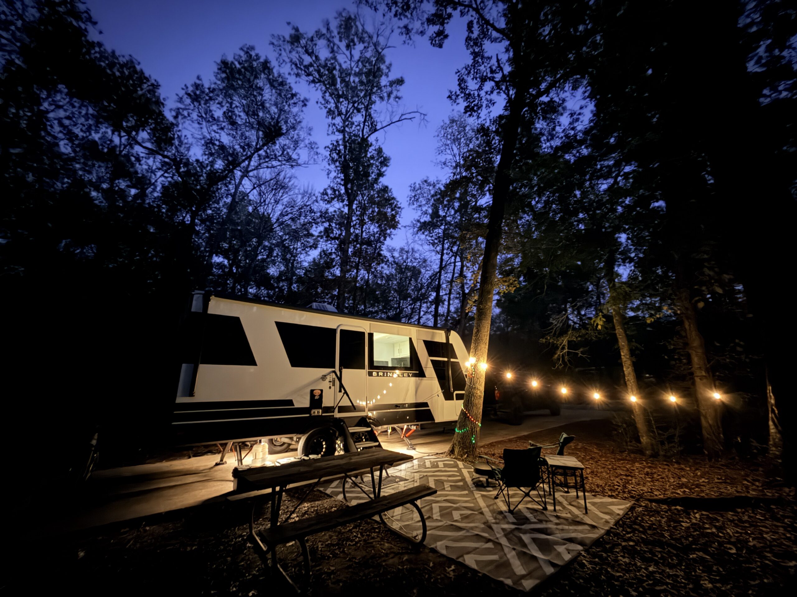 Brinkley trailer glowing at night at a wooded Texas state park campsite with string lights, rug and picnic table.