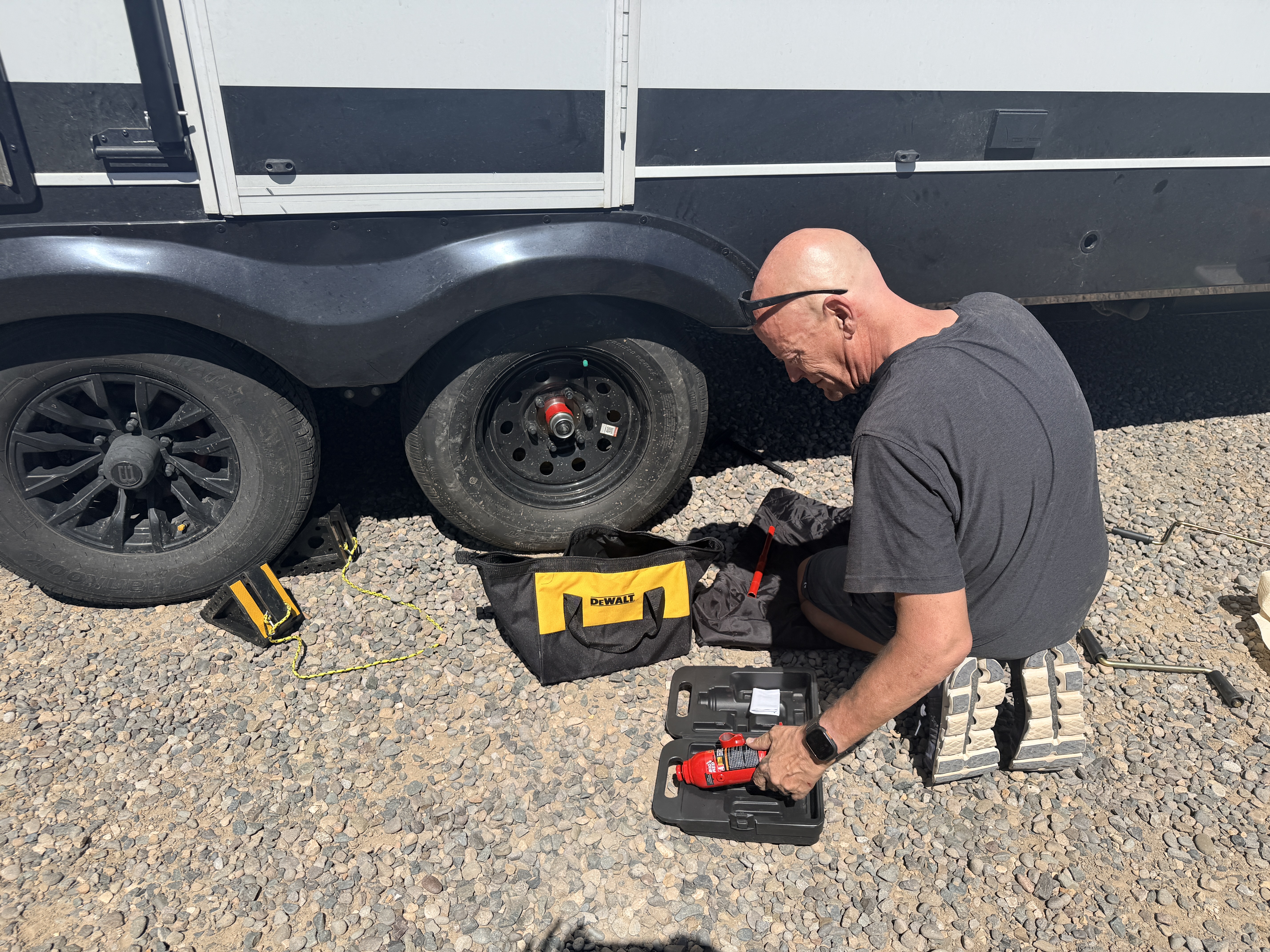 Matt kneeling on gravel with a bottle jack and tool bag, working on a tire of the travel trailer.