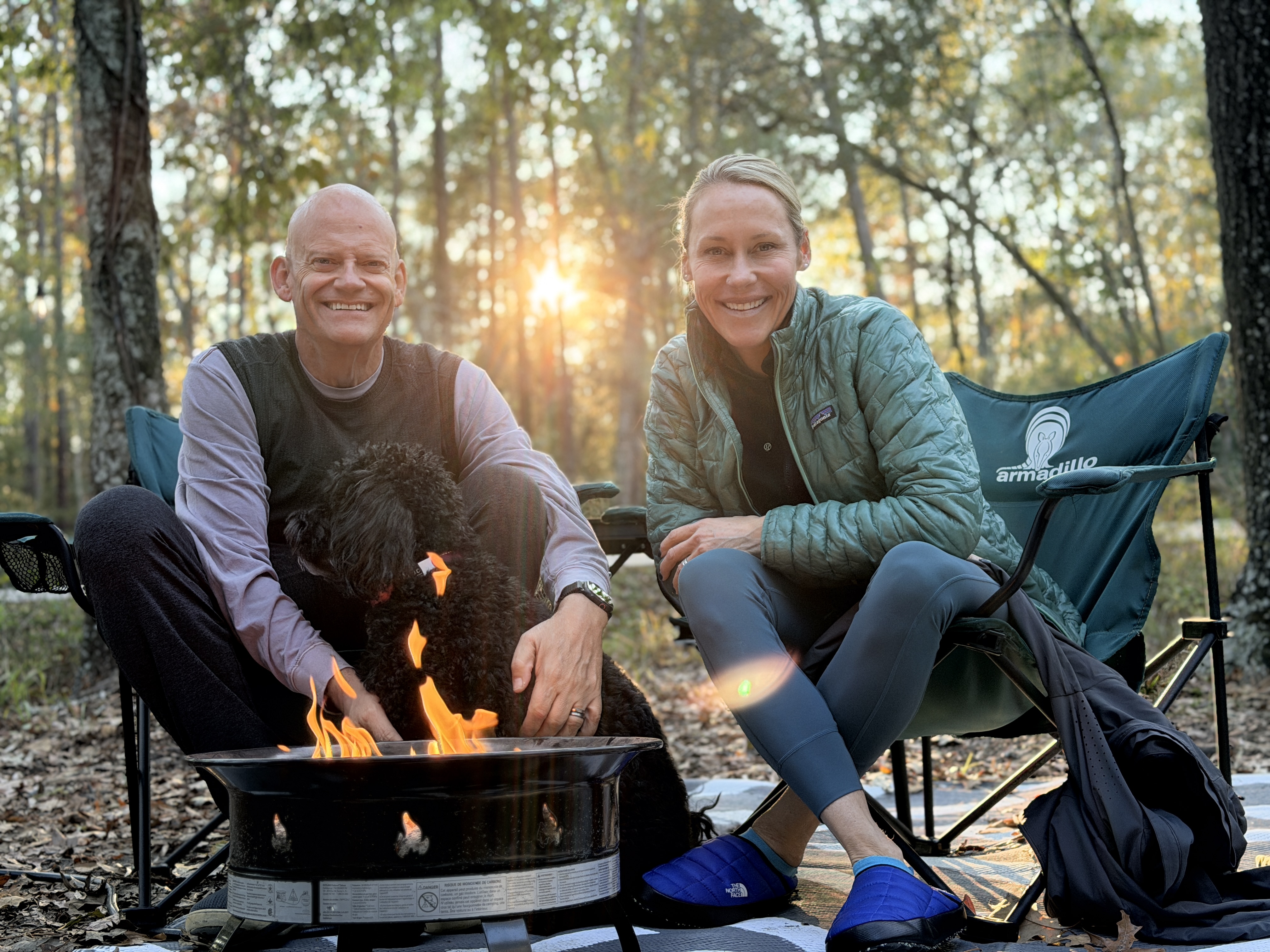 Matt and Erin smiling in camp chairs with Rindo beside a propane fire pit as the sun sets through the pines.